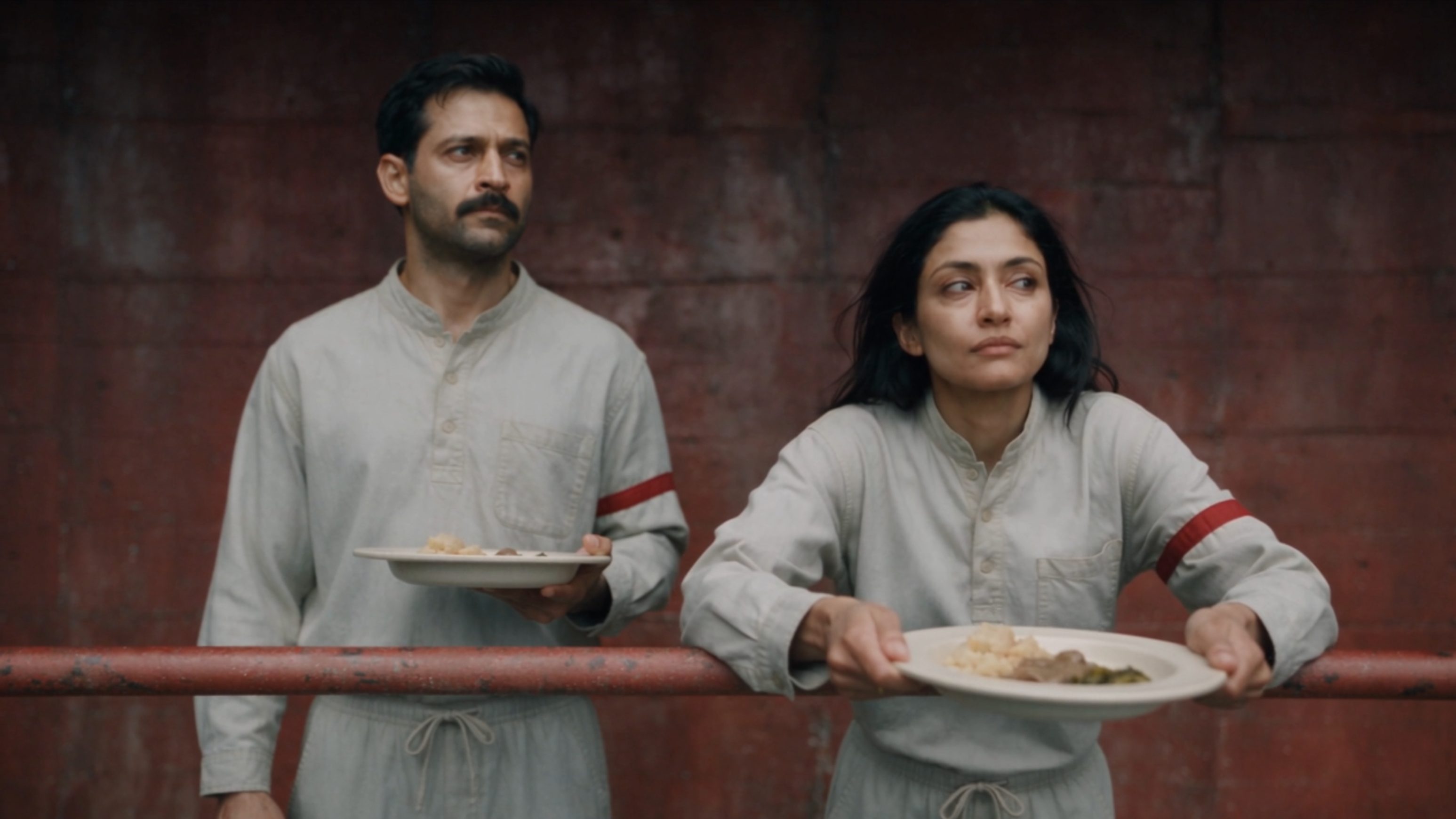 Two actors in grey uniforms hold plates against a rusted red wall